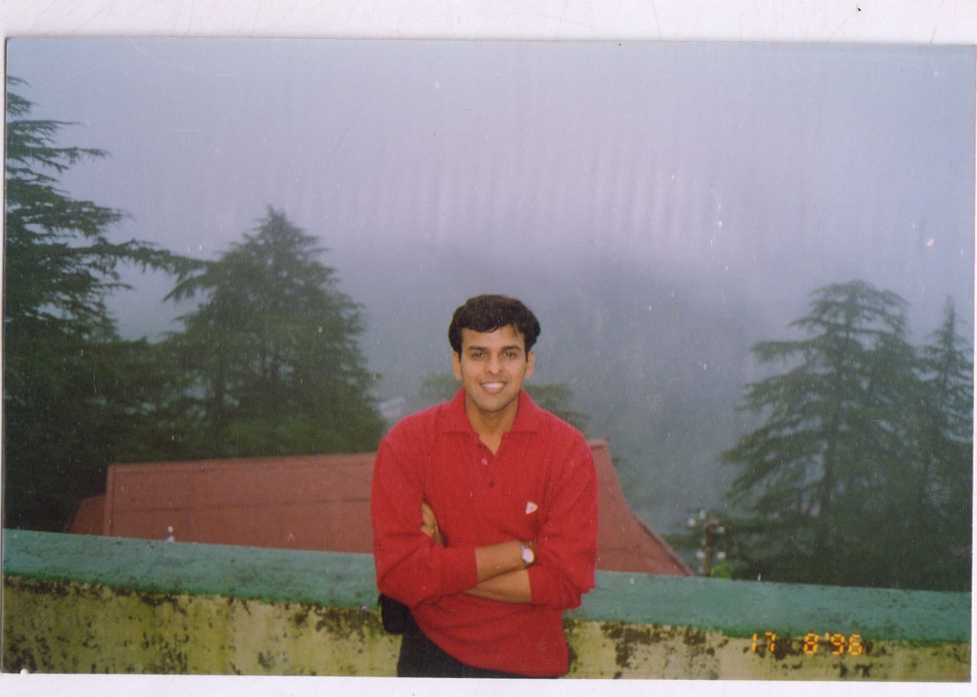 Manuj Aggarwal as a boy, in a red shirt with his arms folded, standing at a low wall on a misted hillside among deodar trees.