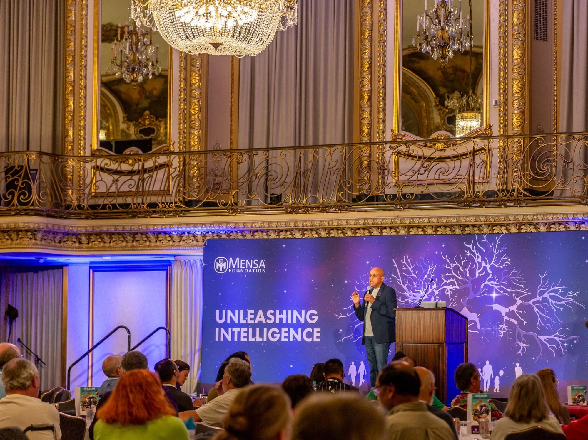 Manuj Aggarwal speaking on stage at a Mensa Foundation event in a chandeliered ballroom, beneath a backdrop reading Unleashing Intelligence.