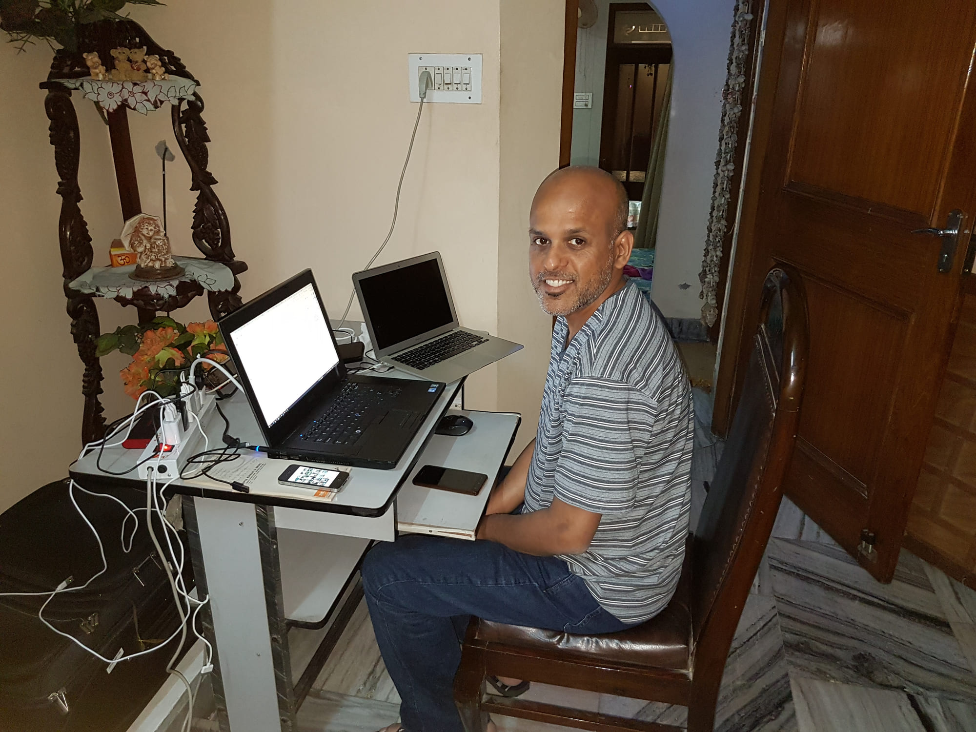 Manuj Aggarwal seated at a small folding desk with two laptops, in a room with carved wooden furniture and a marble floor.