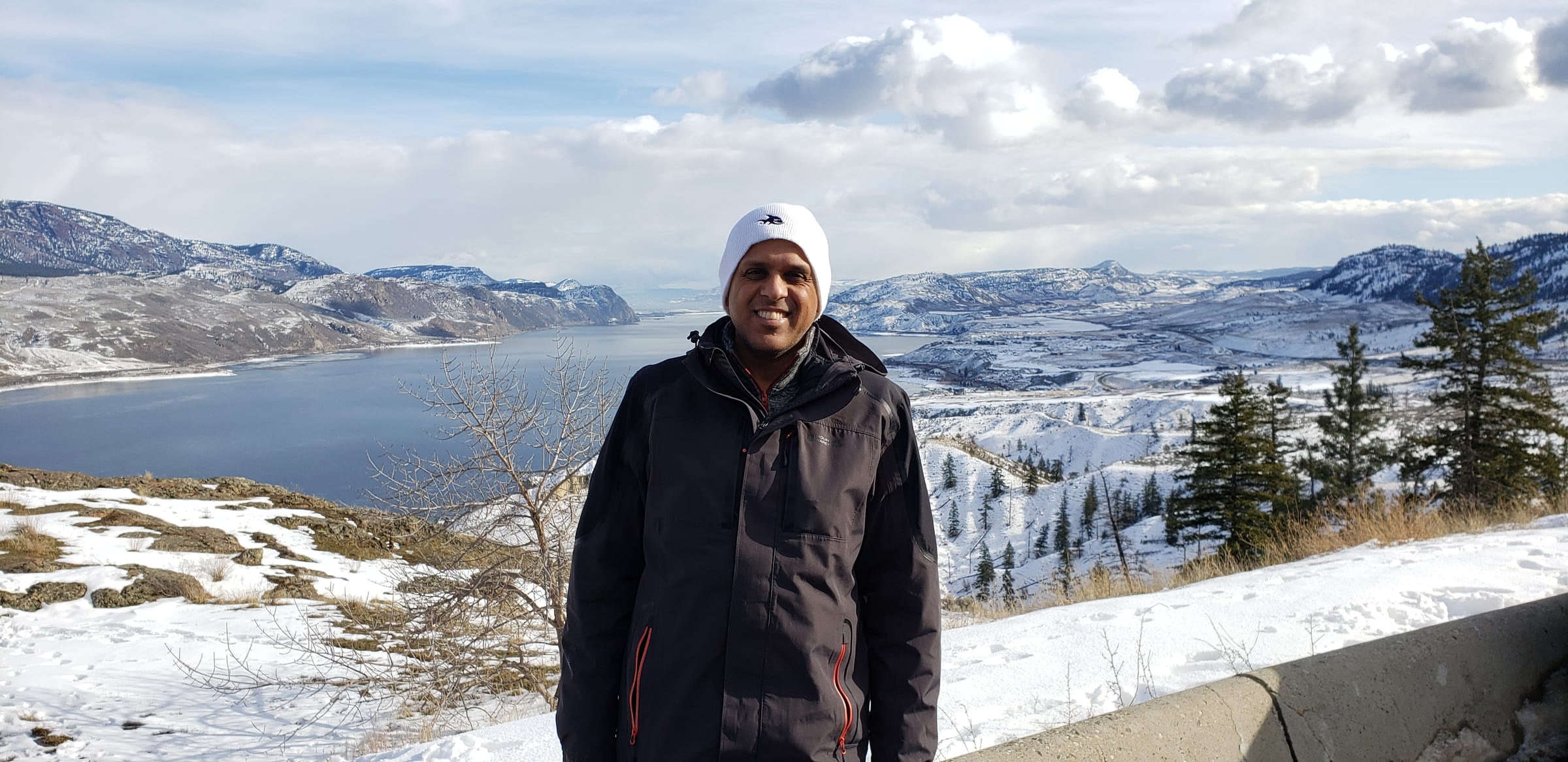 Manuj Aggarwal in a winter coat and white hat, standing on a snowy ridge above a long lake, with snow-covered hills stretching to the horizon.